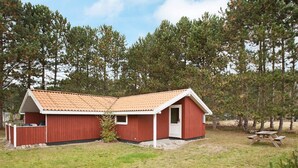 Une cabane en bois rouge avec un toit en tuiles, un petit porche et une table de pique-nique, située dans un espace verdoyant, avec des pins en arrière-plan.