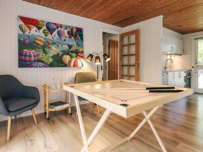 A cozy room with a wooden ceiling, a large painting of hot air balloons, a table with a board game, a chair, and a small side table with books.