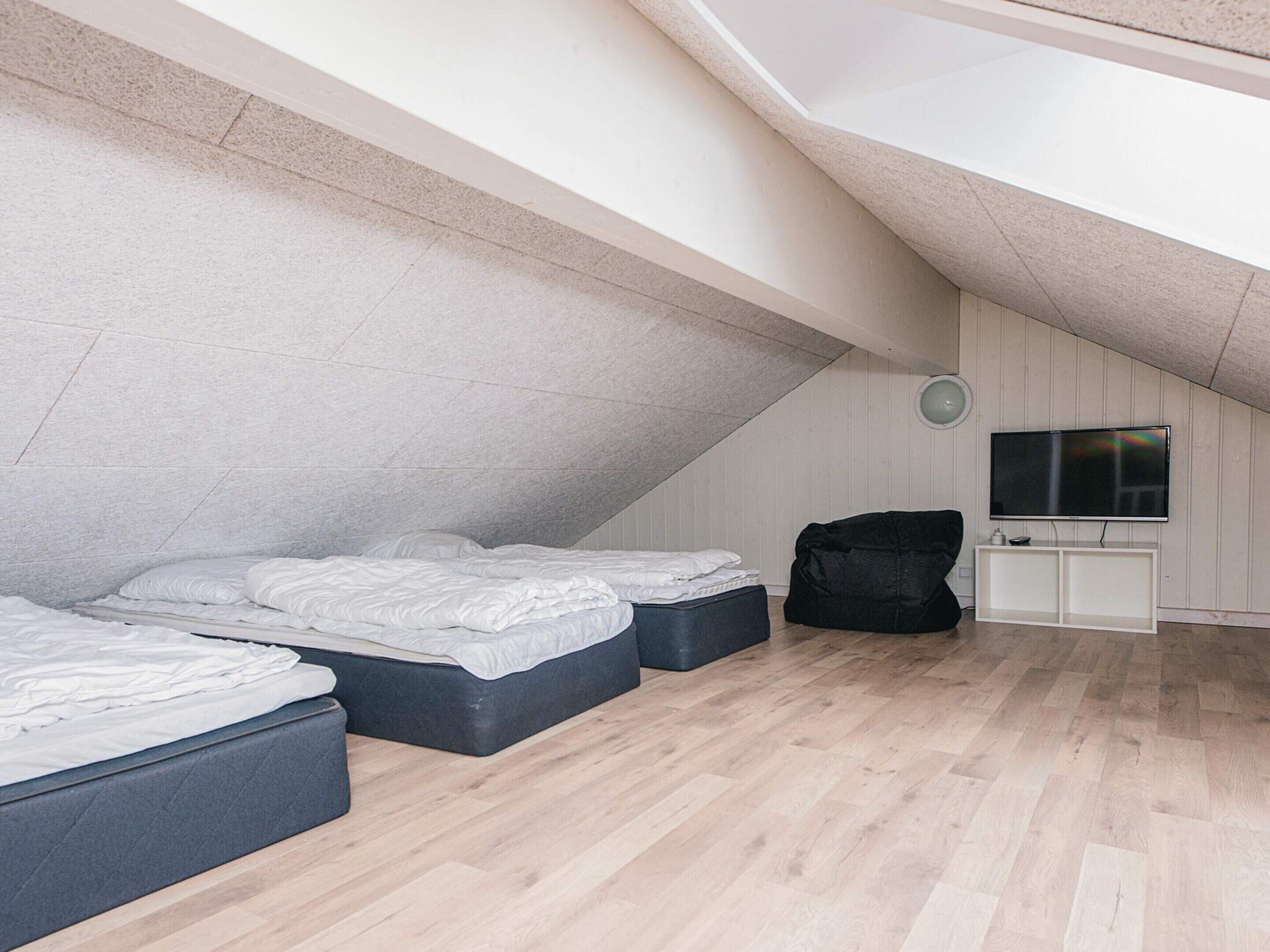 Une chambre mansardée avec un plafond haut et une pente, deux lits, un fauteuil en forme de haricot noir, un téléviseur posé sur un meuble blanc et un plancher en bois.