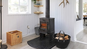 A cozy room with a wood-burning stove, white paneled walls, a window, and a wicker basket. - Tranquil Retreat in Blavand - By Traum Ferienwohnungen (Blavand)