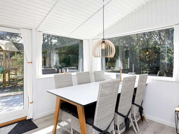 A dining area with a white table and wicker chairs, large windows, and a hanging light fixture.