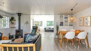 A cozy living room with a sofa, a dining table with chairs, a fireplace, and a view through an open door to a balcony. - Tranquil Retreat in Blavand - By Traum Ferienwohnungen (Blavand)