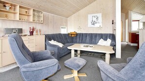 Een modern interieur met een houten plafond, een grijze zithoek, een kleine tafel en een keuken met houten kastjes en een aanrecht.