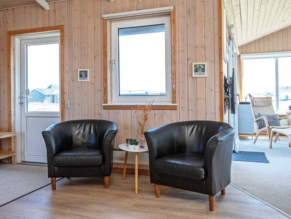 A room with two leather chairs, a small table with a vase, wooden flooring, and large windows with wooden frames.