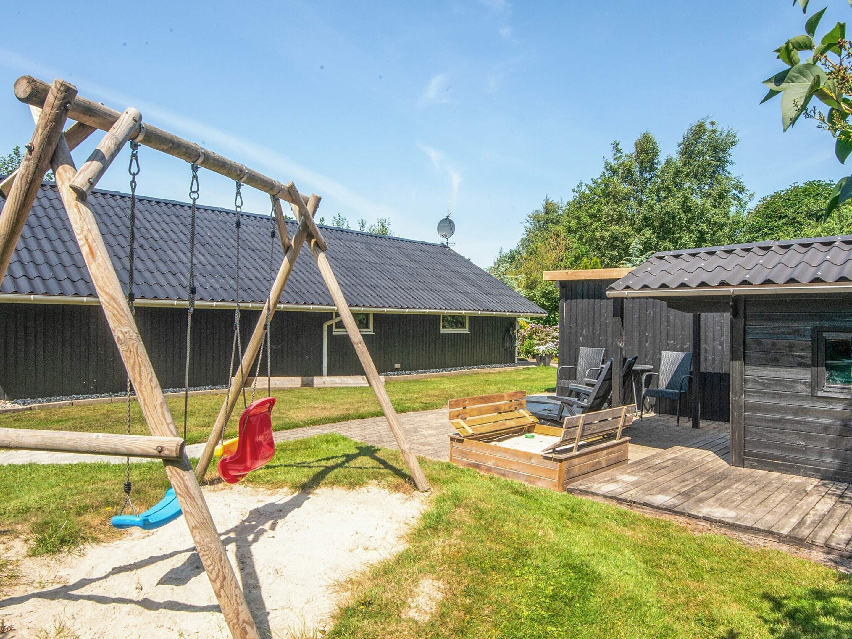 A wooden swing set, a small deck with seating, and a building with a dark roof.