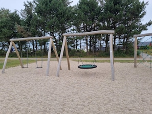 A playground with two swings and a tire swing on a sandy surface, surrounded by trees and a fence.