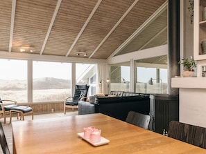 A spacious room with a wooden ceiling, large windows, a black leather sofa, a dining table with pink candles, and a fireplace. - 4 Star Holiday Home in Lokken (Lokken)
