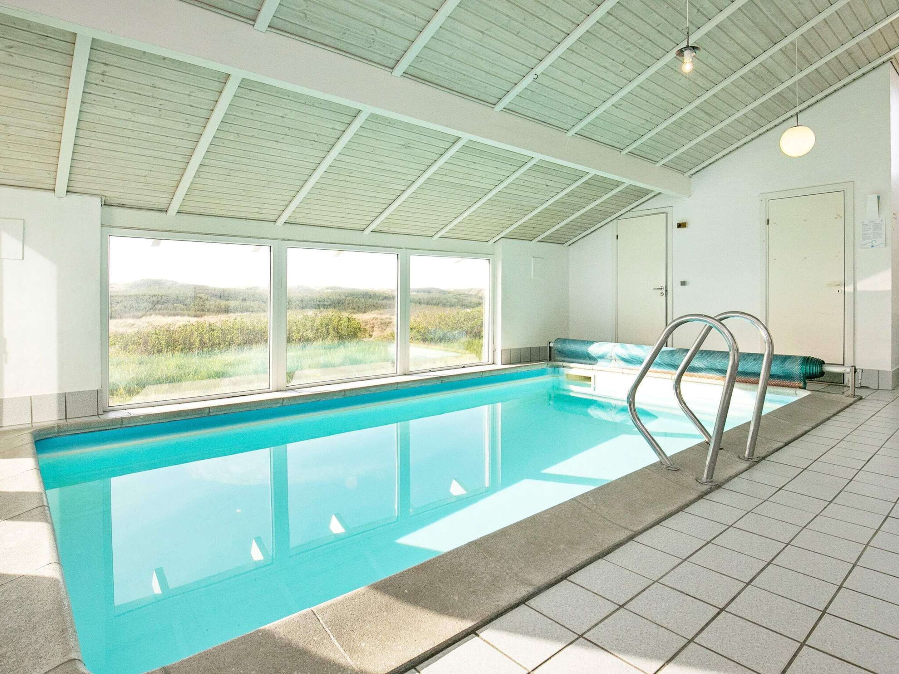 Una piscina interior con ventanas transparentes, suelo de baldosas y escalera metálica.