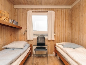 A small room with two beds, a chair, a shelf with books, and a window with curtains. - 4 Star Holiday Home in Lokken (Lokken)