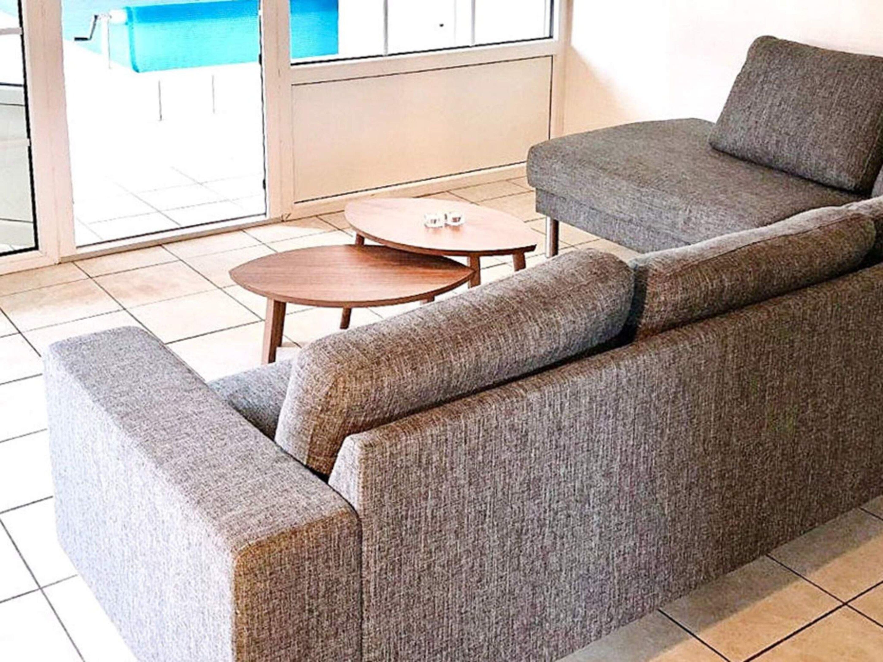 A grey sofa with a wooden side table and a small round table in a room with tiled flooring.