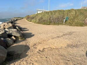 A sandy pathway beside a rocky coastline with a grassy embankment and a modern building structure.