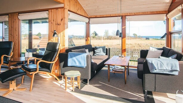A spacious room with a wooden ceiling, large windows, and a view of the beach. There are several seating areas with chairs and a sofa. - 4 Star Holiday Home in Juelsminde (Juelsminde)