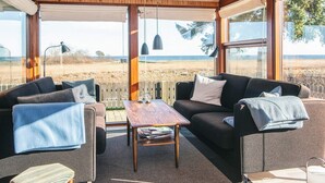 A cozy room with a wooden ceiling, large windows, a sofa, a table, and a view of the beach. - 4 Star Holiday Home in Juelsminde (Juelsminde)