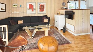 Ein gemütliches Wohnzimmer mit einer schwarzen Couch, einem Holztisch, einem Lederhocker und einem Teppich. Es gibt eine Küchenzeile mit einem weißen Schrank und einem Fernseher.