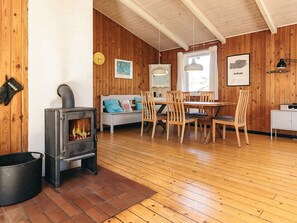Ein gemütliches Blockhaus-Interieur mit Holzofen, Essbereich mit Stühlen und Tisch, einer Couch mit Kissen sowie einem Fenster mit Vorhängen.