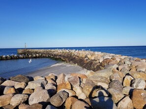 Ein felsiger Wellenbrecher, in der Ferne ein Leuchtturm, ein Sandstrand und Möwen.