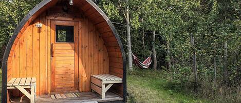 A wooden cabin with a pitched roof, a small window, and a door. There is a bench outside and a hammock in the background.