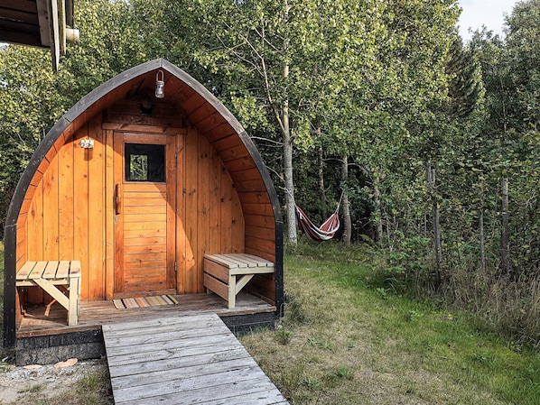 A wooden cabin with a pitched roof, a small window, and a door. There is a bench outside and a hammock in the background.