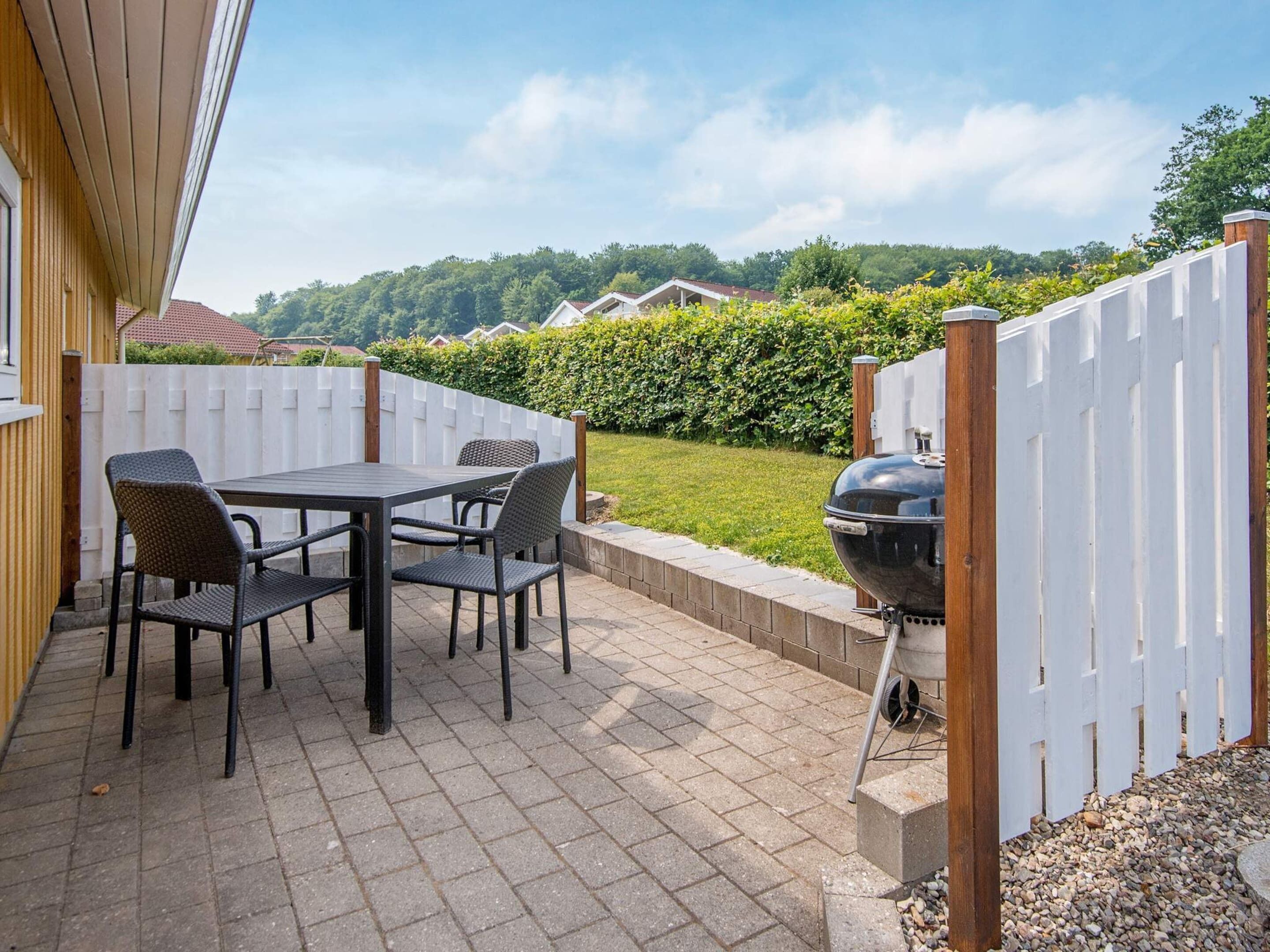 An outdoor patio with a table and chairs, a barbecue grill, and a white fence.