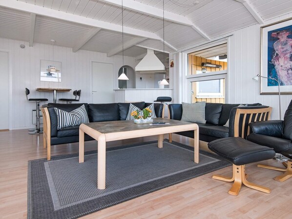 A modern living room with a sofa, coffee table, and chairs. There is a kitchen area visible through an open door. - 4 Star Holiday Home in Sjolund (Sjølund)