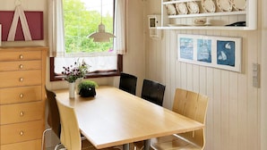 A dining area with a wooden table, four chairs, a vase with flowers, a window with curtains, and a shelf with plates and a framed picture. - 5 Person Holiday Home in Lemvig (Lemvig)