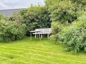A grassy area with a white bench and a table, surrounded by dense foliage and a building with a tiled roof in the background.