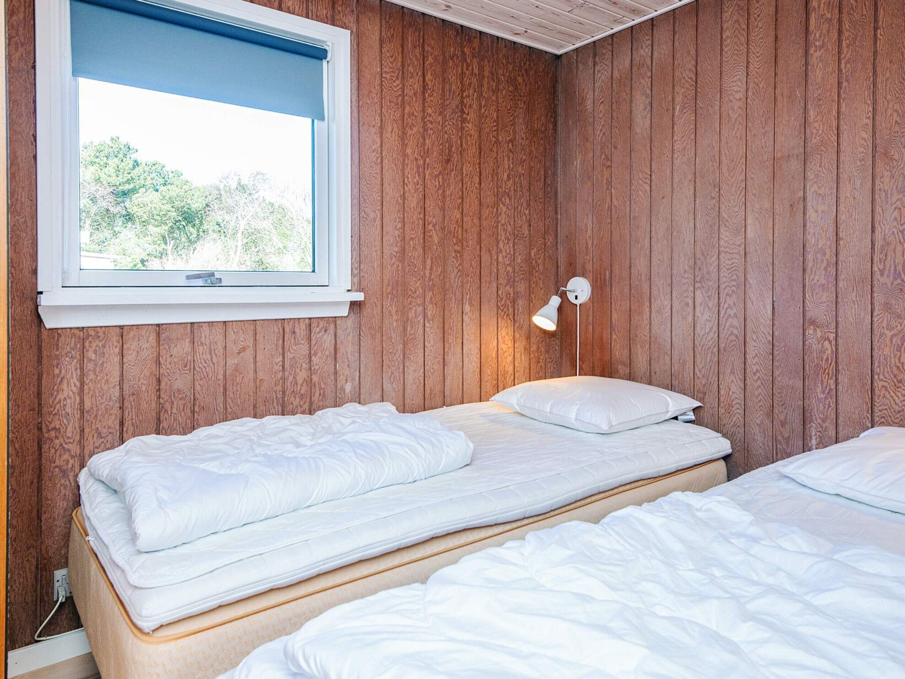 A room with wooden panel walls, a window with a view of trees, a wall-mounted lamp, and two beds with white bedding.