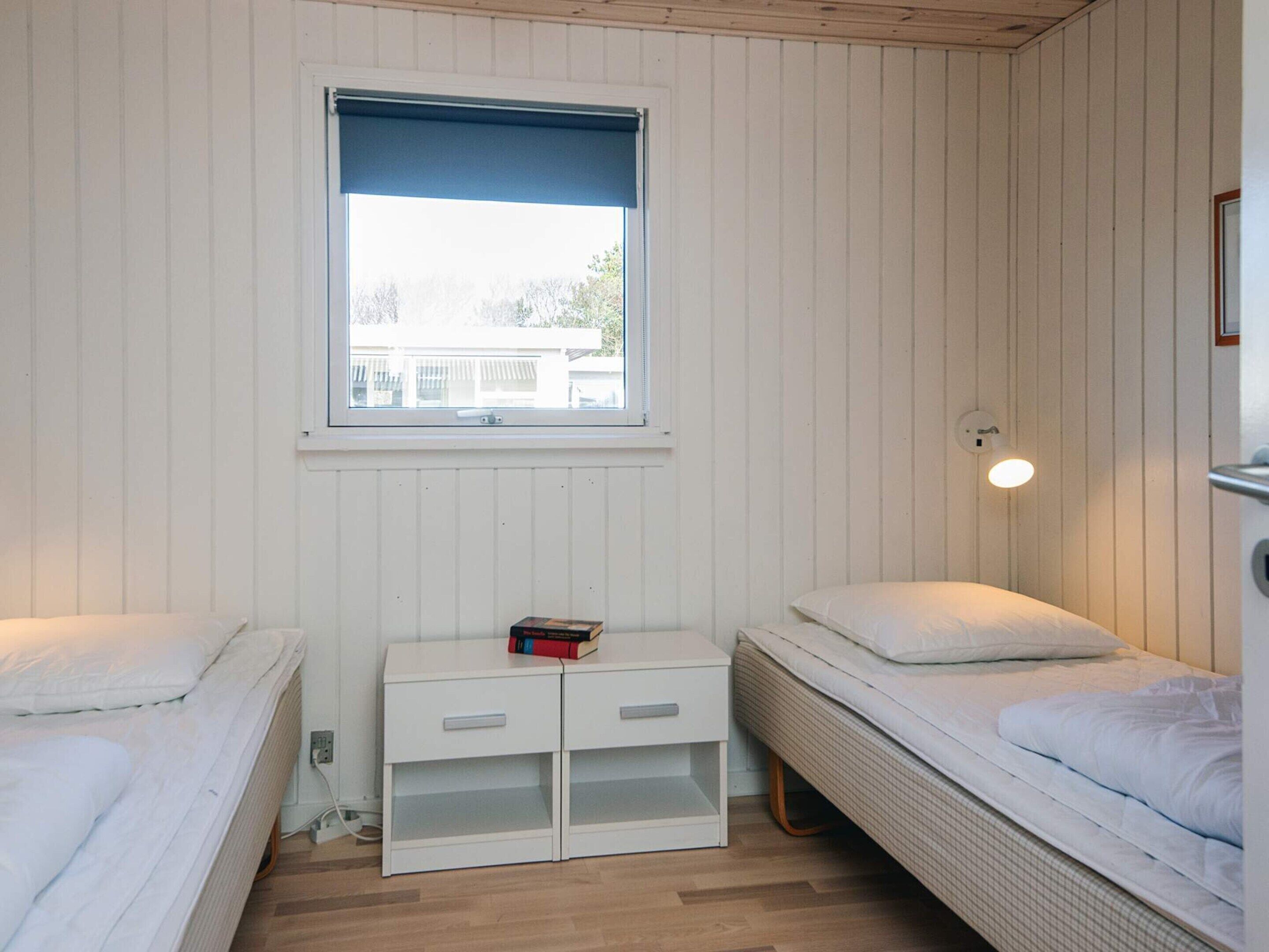 A small, single-room accommodation with two beds, a window, a bedside table with a book, and a wall-mounted light.