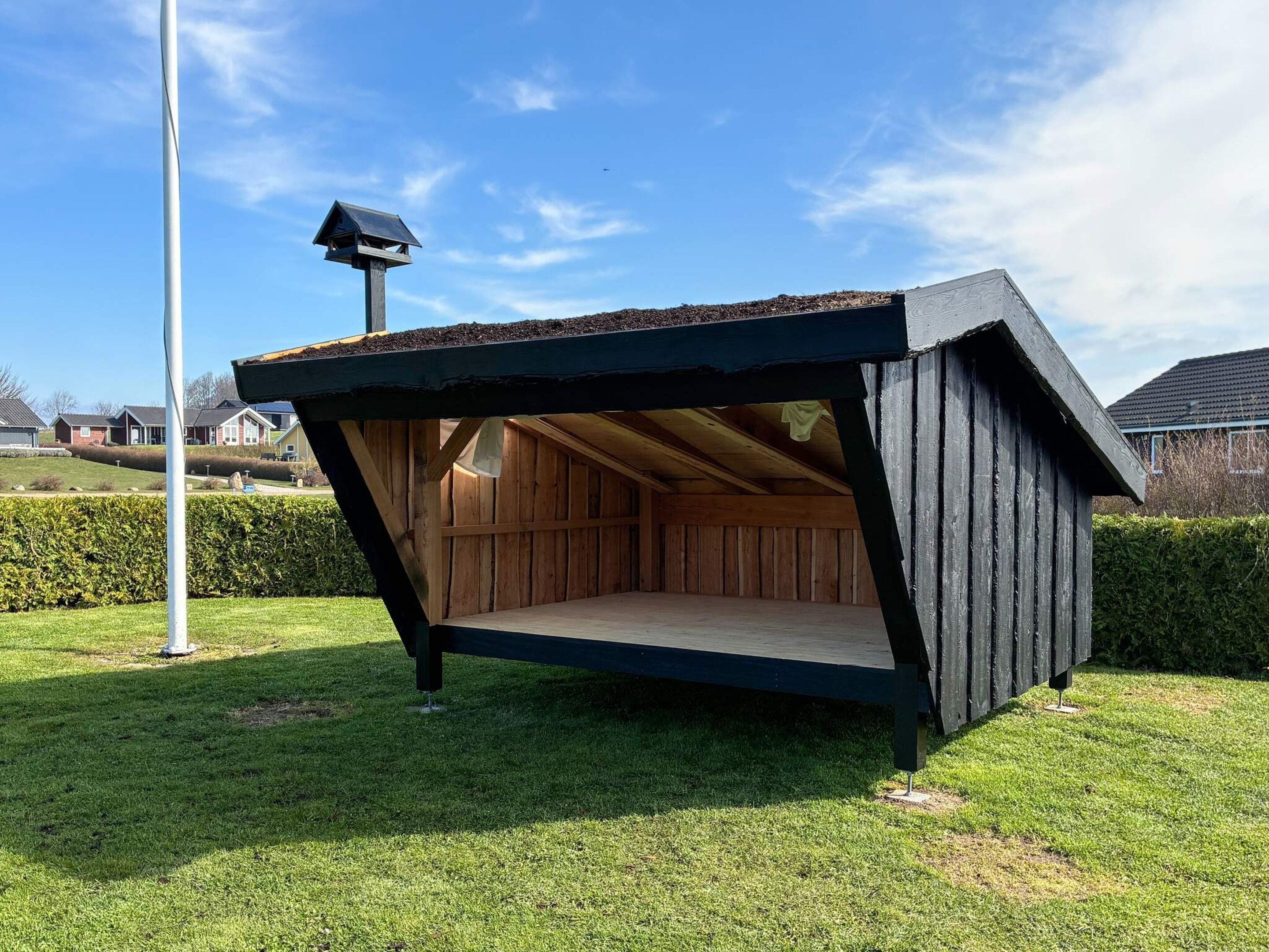 A wooden structure with a slanted roof, possibly a shed or storage unit, in a grassy area with a clear sky.
