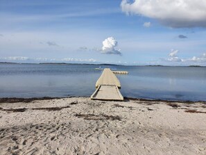A sandy beach with a wooden pier extending into calm water, surrounded by a clear sky.