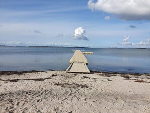 A concrete pier extending into calm water, surrounded by sandy beach with visible footprints.