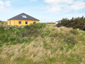 Een geel huis met een donker dak, omringd door hoog gras en struiken.