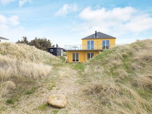 A yellow house with a balcony, surrounded by grass and a rocky path. - 5 Star Holiday Home in Brovst (Brovst)