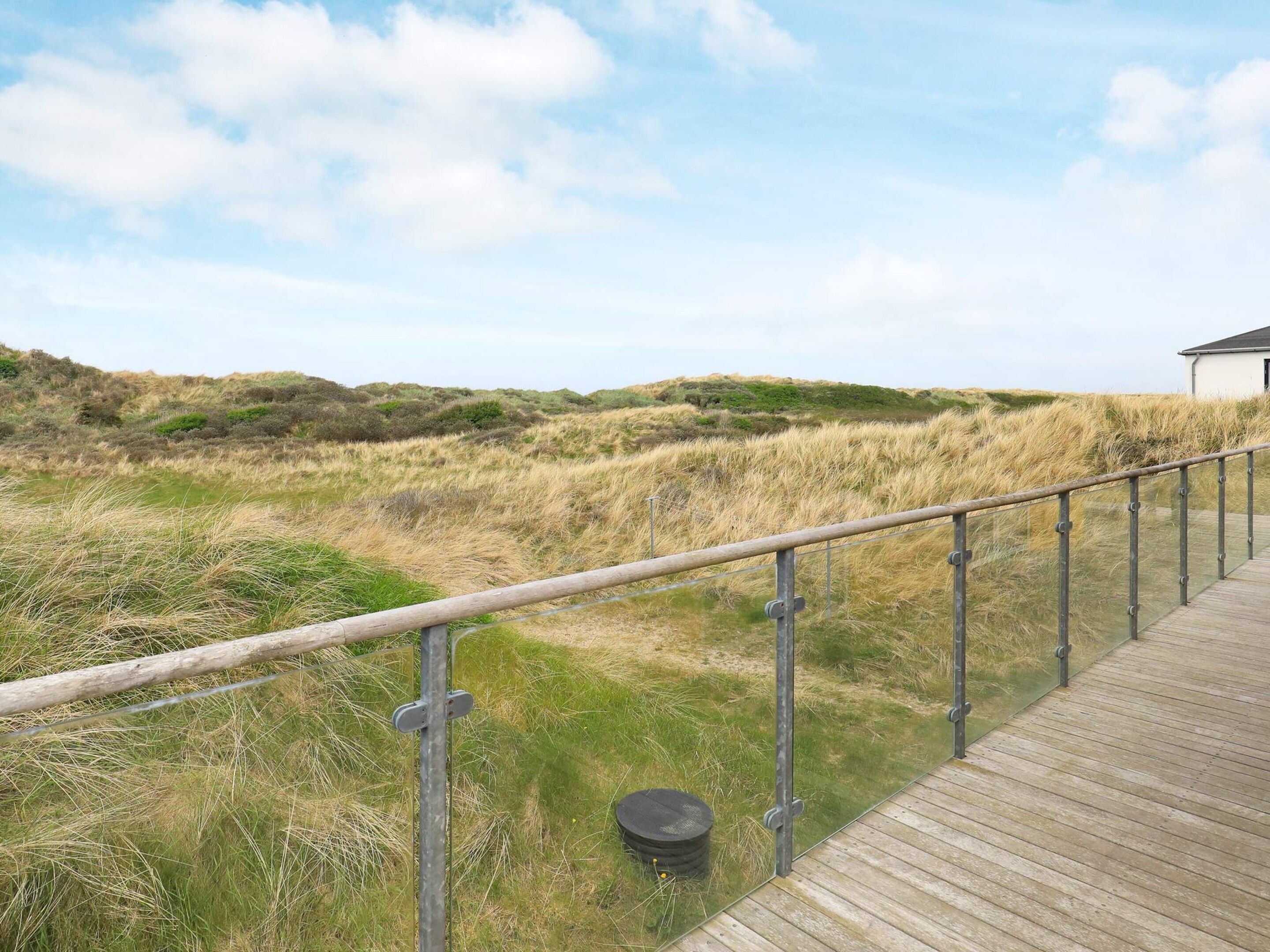 A wooden boardwalk leading to a dune landscape with a grassy field and a building in the distance.