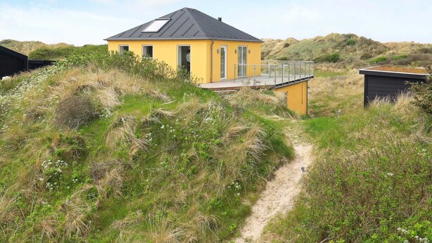 En gul bygning med mørkt tag, omgivet af naturlig vegetation og en sandsti.