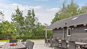 An outdoor patio area with a wooden table and wicker chairs, surrounded by a brick floor and a wooden building with a tiled roof. - Holiday Home in Kappeln (Kappeln)