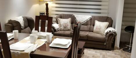 A dining area with a wooden table and chairs, a sofa set, a lamp, and a clock on the wall.