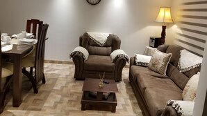 A living room with a brown sofa, a dining table with chairs, a small coffee table, a clock on the wall, and a lamp. - Nice Apartment 5 Minutes Airport (Bogotá)