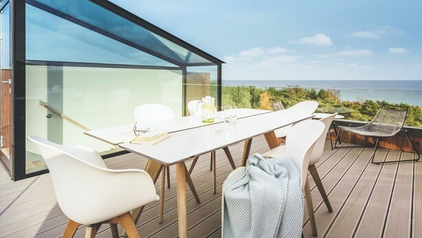 A modern outdoor dining area with a glass enclosure, white chairs, and a view of the sea. - Beautiful apartment for 2 people with WIFI, pool, TV, terrace and pets allowed (Sassnitz)