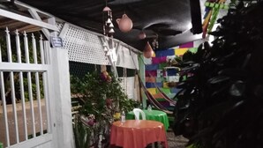 Uma área de pátio externo com uma mesa coberta por uma toalha vermelha, cercada por plantas em vasos e um portão branco.