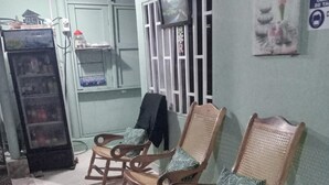 Quarto com três cadeiras de balanço de madeira com almofadas estampadas, um refrigerador e uma TV fixada na parede.
