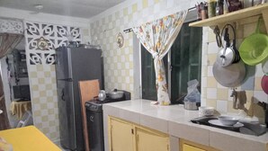 Uma cozinha com fogão, geladeira e diversos utensílios. Há uma mesa amarela com uma cesta de ovos e legumes.