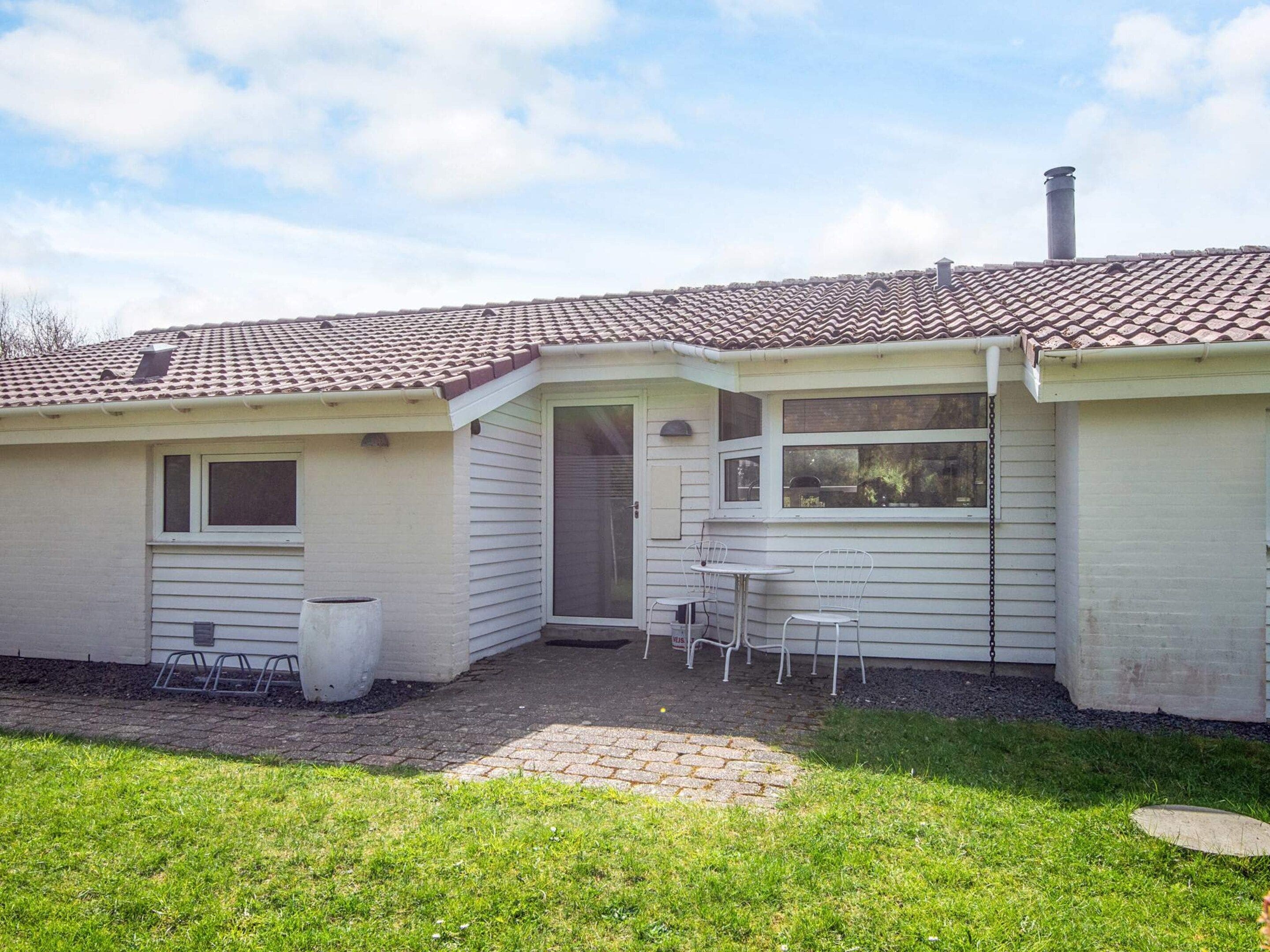 A single-story house with a tiled roof, a small porch with two chairs, and a window overlooking the yard.