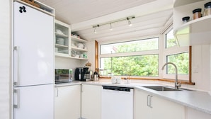 A modern kitchen with white cabinetry, a built-in dishwasher, a coffee maker, and a window providing a view of greenery outside. - 4 Star Holiday Home in Vejers Strand (Vejers Strand)