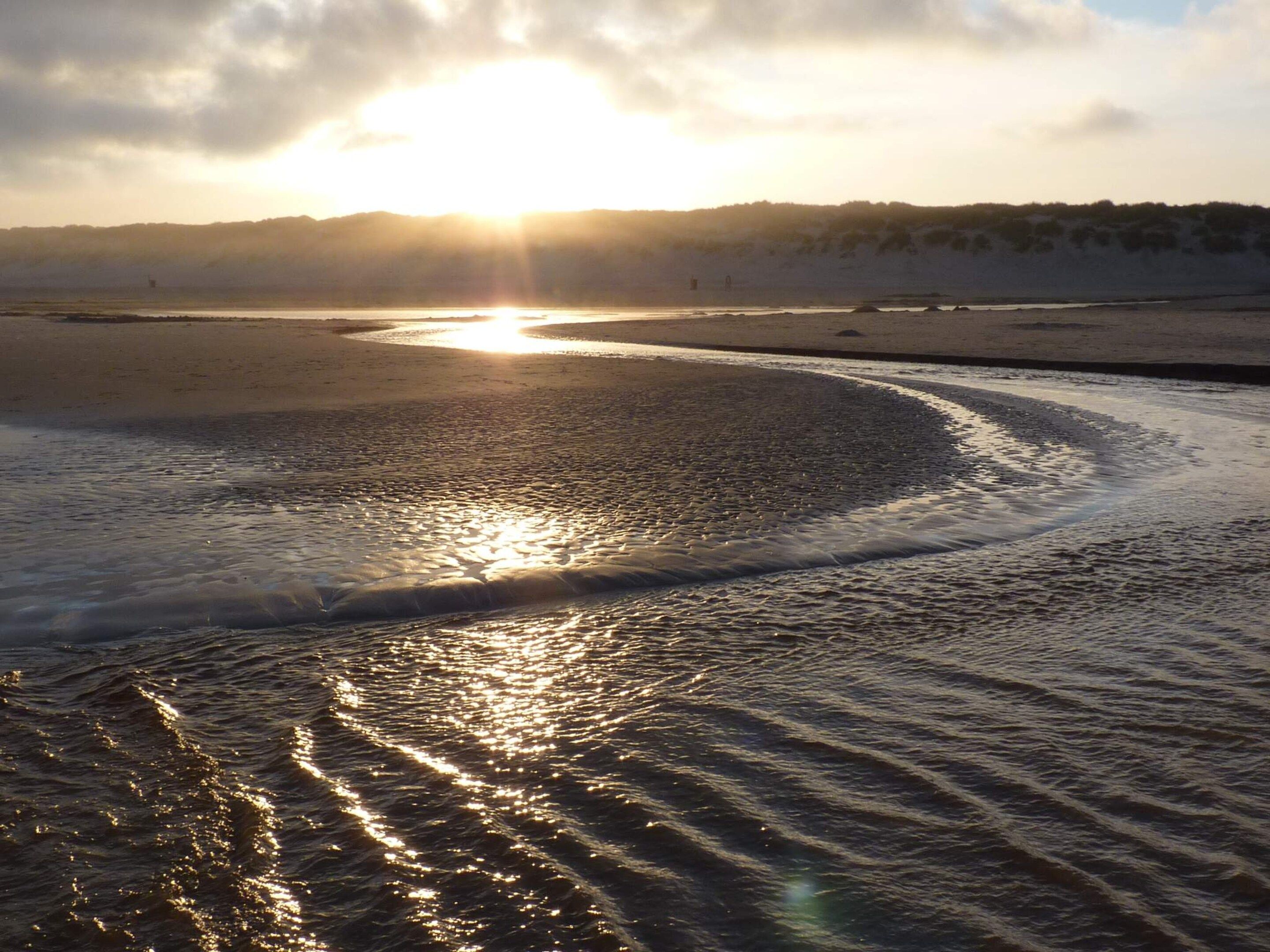 A beach with a calm sea reflecting sunlight, a clear sky, and distant hills.
