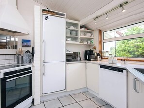 Une cuisine moderne avec des meubles blancs, un four encastré, un réfrigérateur et une fenêtre donnant sur la verdure à l’extérieur.