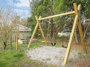 A wooden swing set in a backyard with a sandy ground, surrounded by trees and a shed.