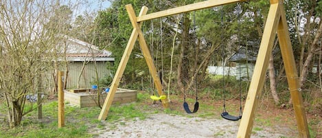 A wooden swing set in a backyard with a sandy ground, surrounded by trees and a shed.
