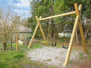 A wooden swing set in a backyard with a sandy ground, surrounded by trees and a shed. - 4 Star Holiday Home in Vaeggerlose (Vaeggerlose)