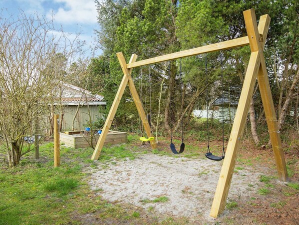 Un portique en bois installé dans un jardin, avec un sol sablonneux, entouré d’arbres et d’un abri de jardin.